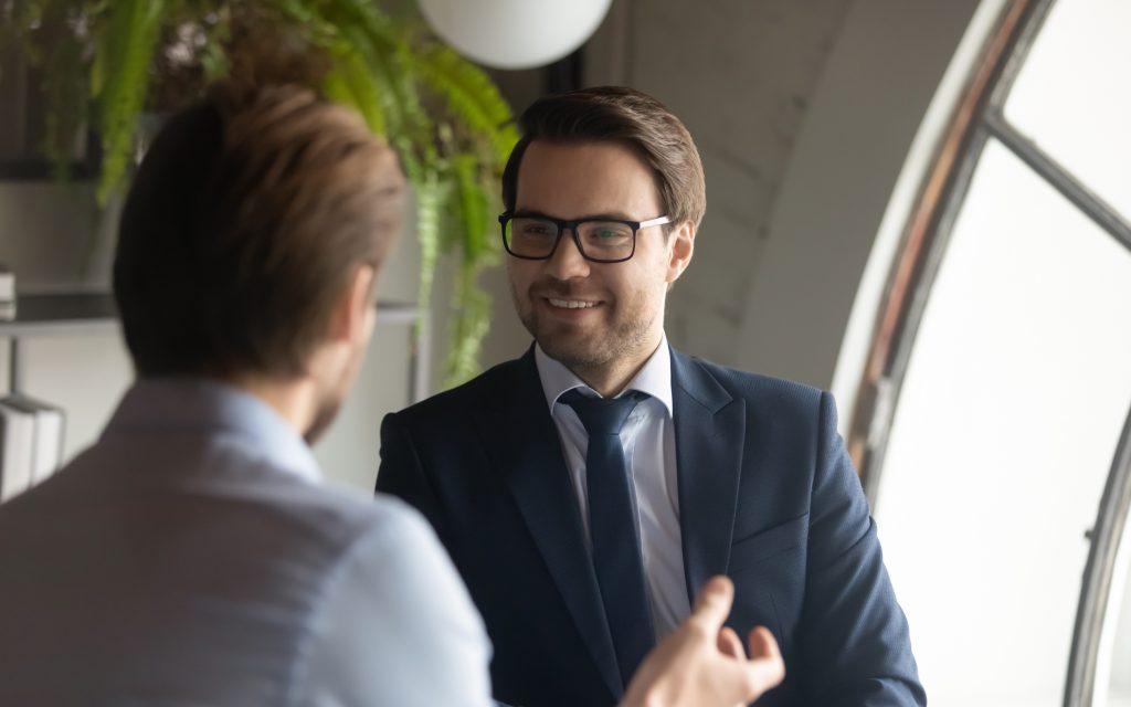 Two people having a conversation, the one in the foreground is facing away from the camera, the one in the background is talking to the person in the foreground.