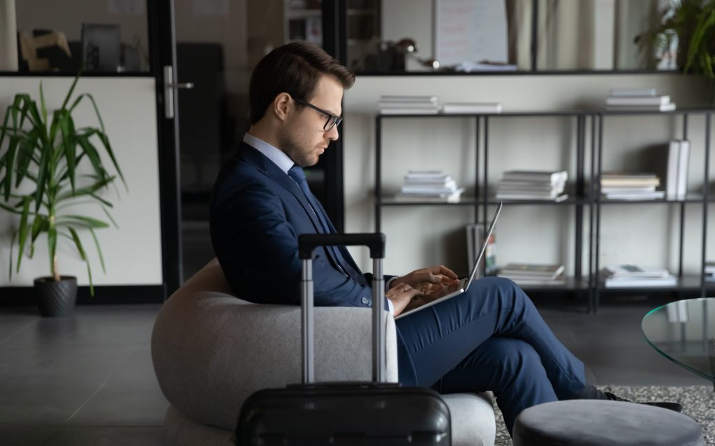 A man getting ready to travel, a suitcase is next to his chair and he is working on his laptop.