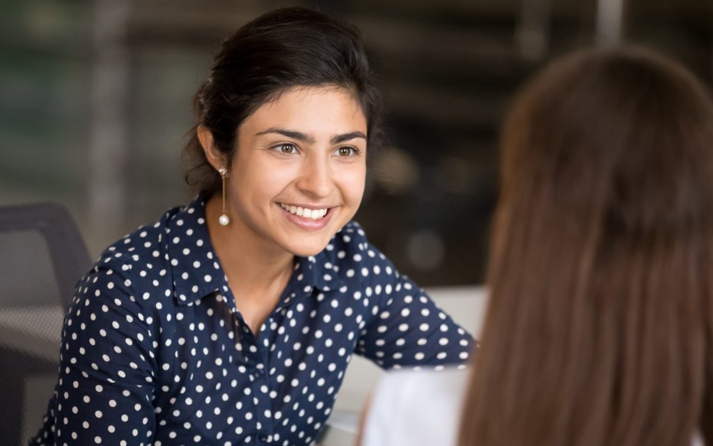 Two people having a conversation. The woman looking towards the camera has a bright smile.